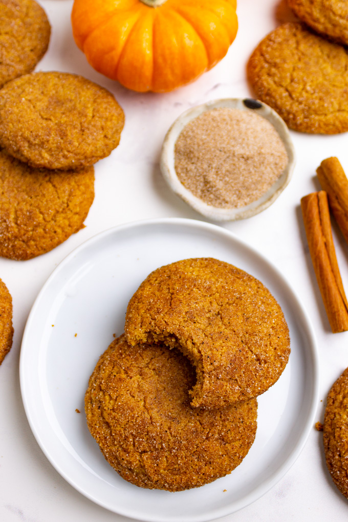 close up side angle of pumpkin snickerdoodle cookies on a plate, one has a bite taken out of it. More cookies, and a pumpkin, and cinnamon sticks are in the background.