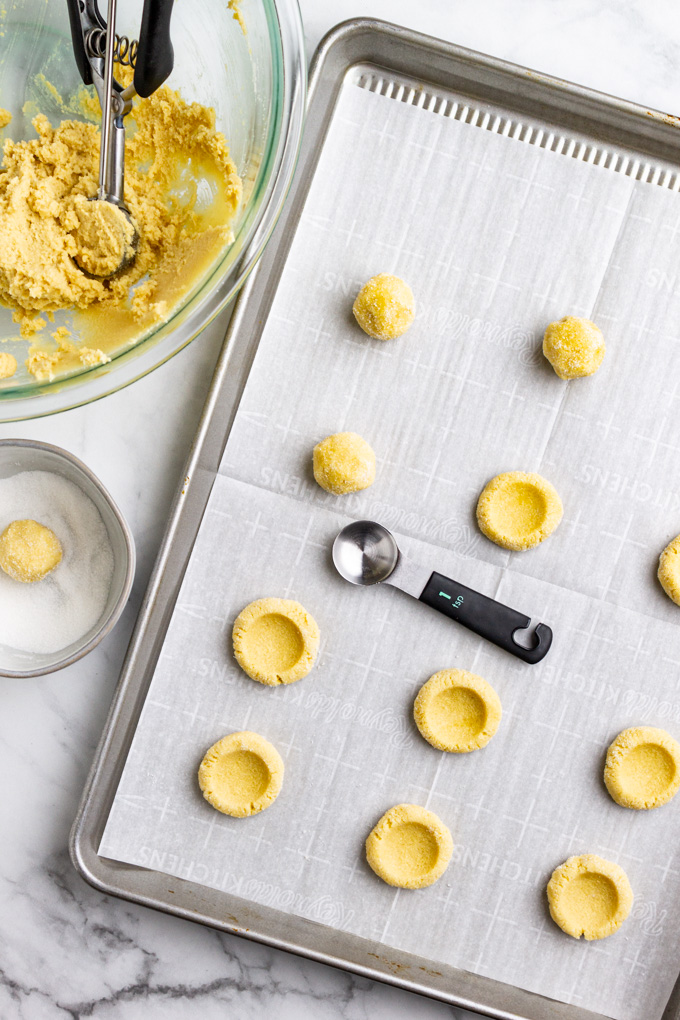 A baking sheet has dough balls of thumbprint cookies with indents made in them.