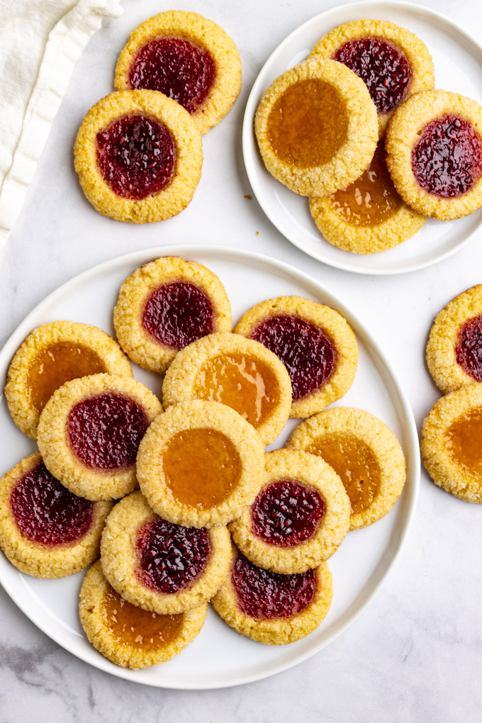 raspberry and apricot thumbprint cookies are stacked on a white plate, on a white background.