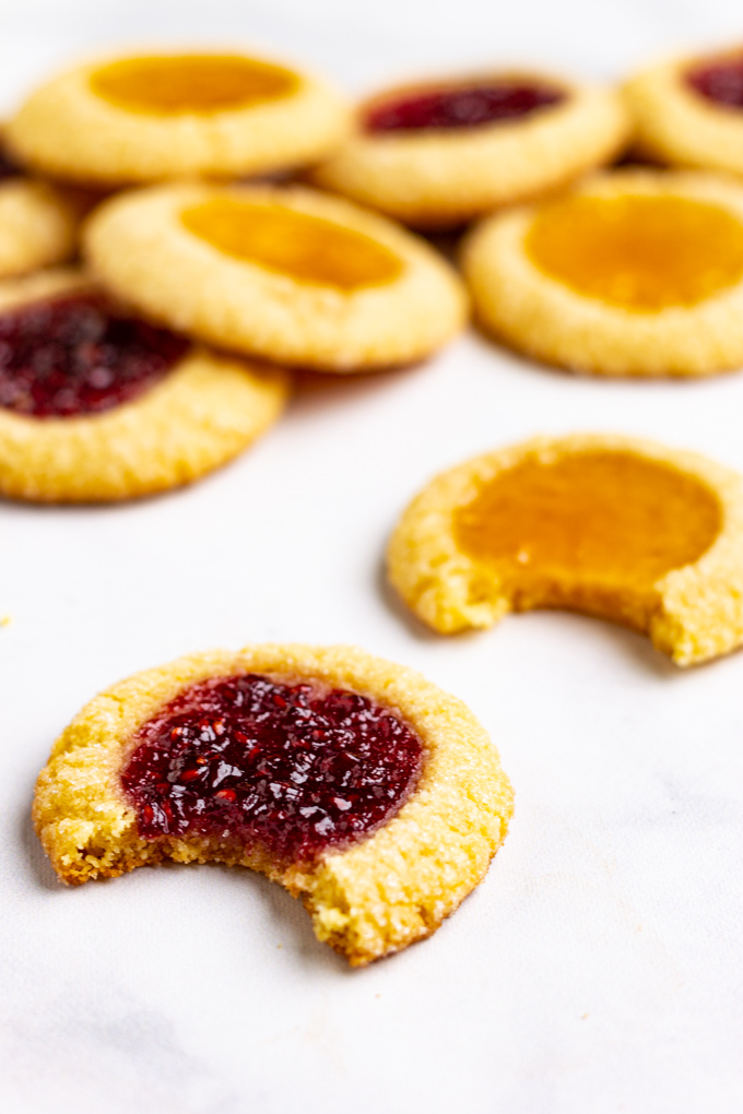 a close up shot of a raspberry thumbprint cookie with a bite taken out of it. A stack of cookies is in the background.