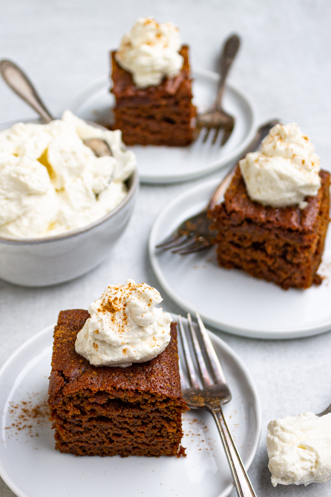 angled shot of gingerbread cake with whipped cream on top, and a bowl of whipped cream in the background.