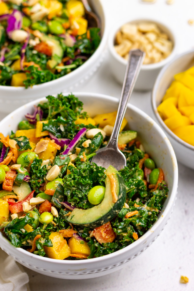 kale Thai salad in a bowl with a fork serving up some of the salad. Mango and peanuts are in separate bowls surrounding it.