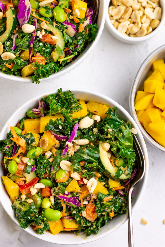 kale Thai salad in white bowls with mango and peanuts in bowls nearby.