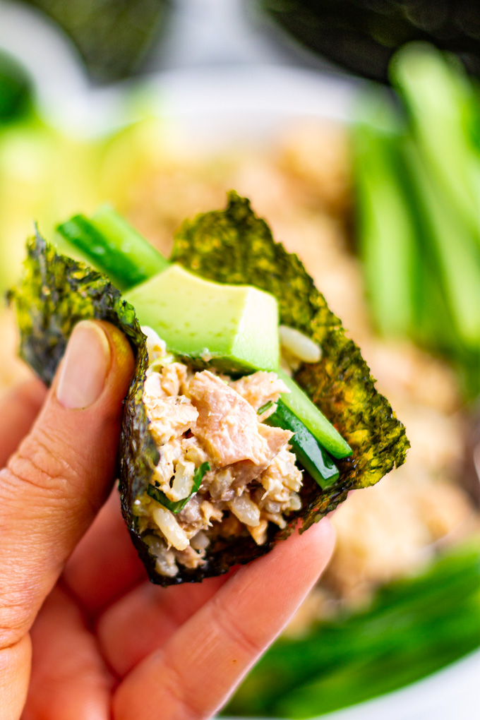 a hand holding a nori sheet that's stuffed with salmon rice bowl along with an avocado slice and cucumbers.