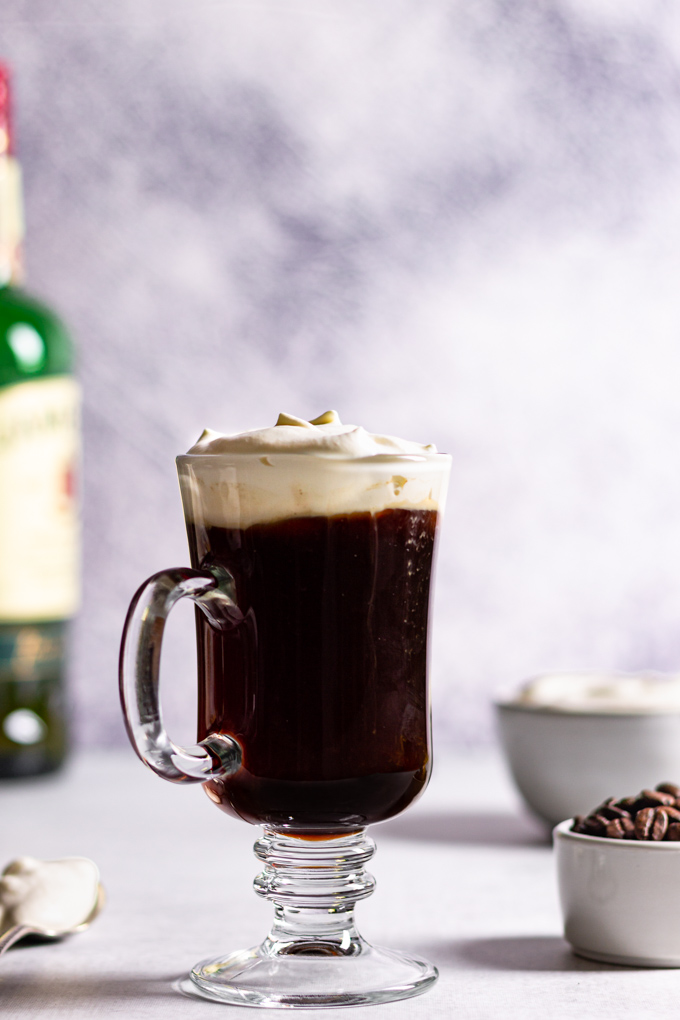 A glass of easy Irish coffee with a layer of coffee and cream on top. There is a bottle of Jameson whiskey in the background. 