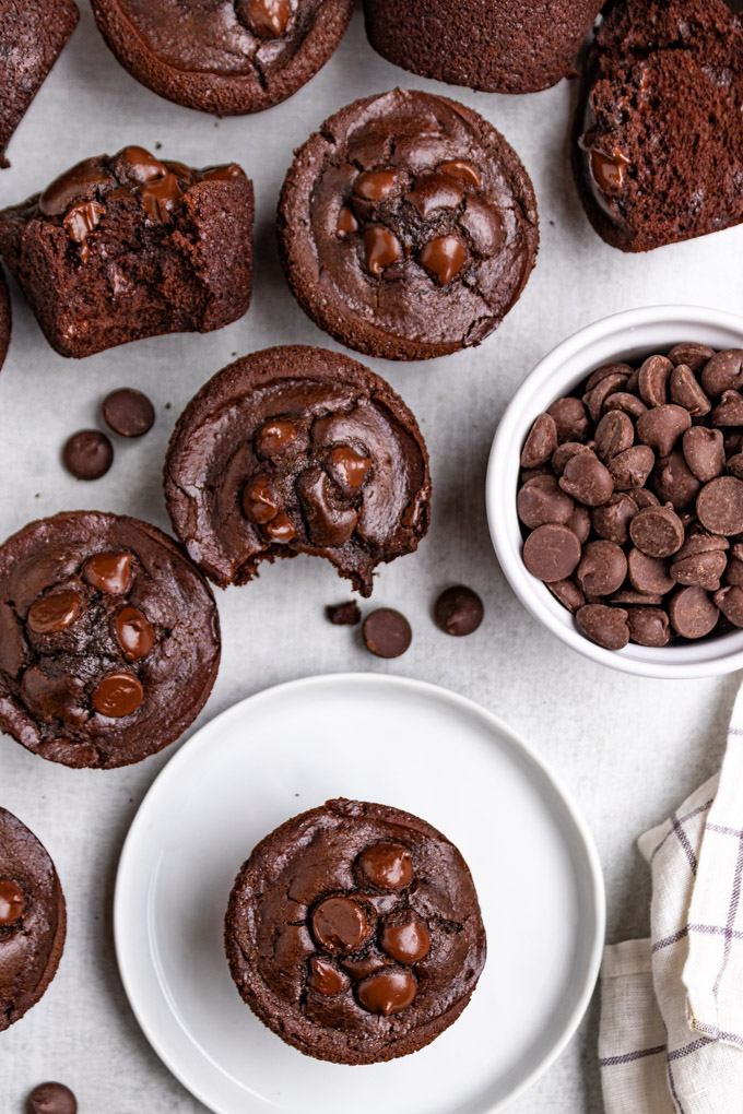 overhead shot of chocolate black bean muffins on a light gray background. One muffin is a on a small white plate. More muffins are scattered in the background. Chocolate chips are in a bowl.