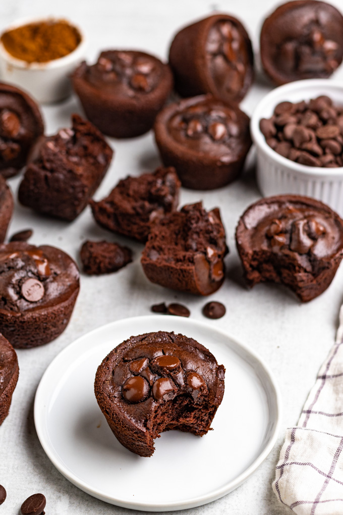 angled shot of chocolate black bean muffins with one muffin on a plate with a bite taken out of it. More muffins are scattered in the background along with a bowl of chocolate chips, and cocoa powder.