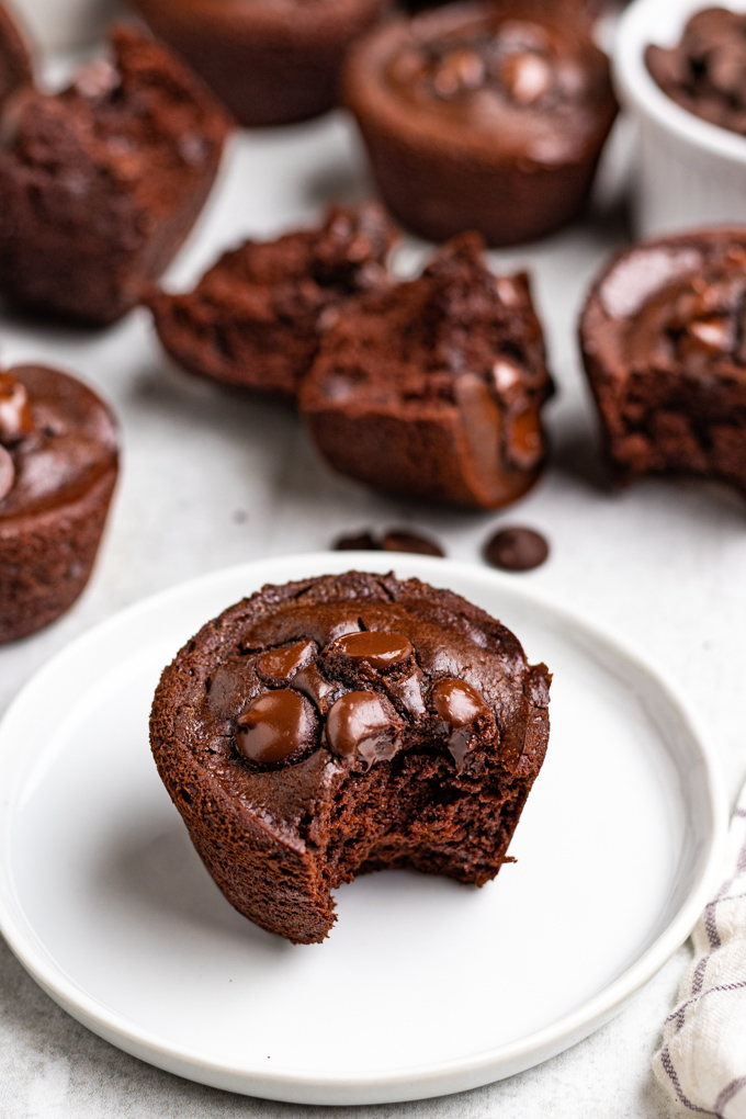 chocolate black bean muffins. One is on a white plate, with a bite taken out of it. More muffins are scattered in the background.