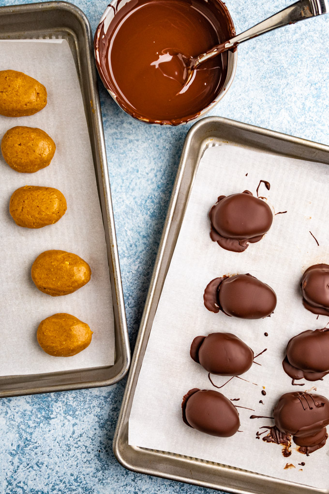 peanut butter eggs on 2 sheet pans, one pan has eggs covered in chocolate, the other pan, the eggs aren't covered in chocolate. There is a bowl of chocolate next to them.