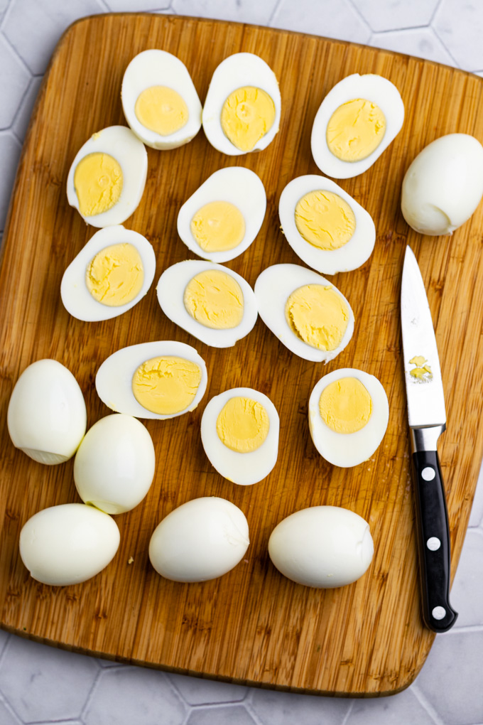 hard boiled eggs on a cutting board, some are sliced in half lengthwise, and others are still whole. A pairing knife is on a corner of the cutting board.