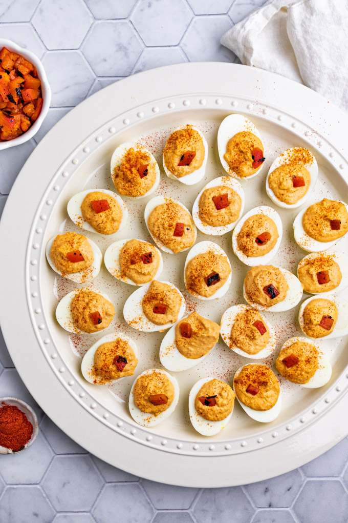 Overhead shot of roasted red pepper deviled eggs on a large white platter. There is a bowl of roasted red peppers, and smoked paprika off to the sides.