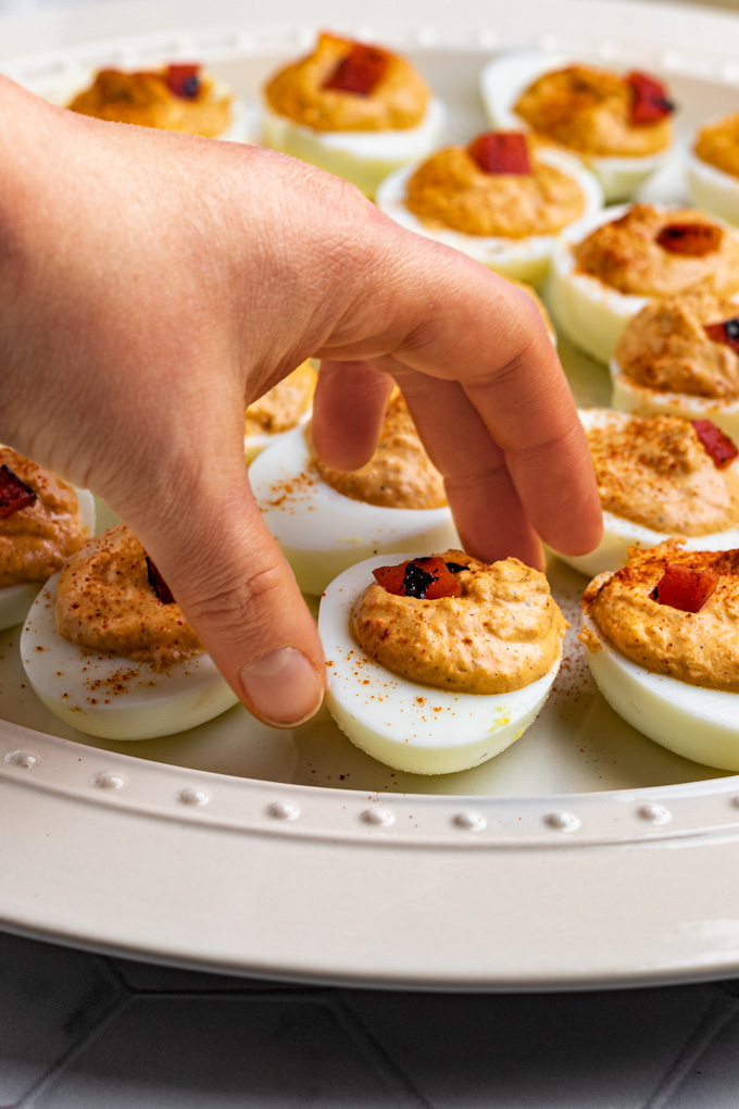 A close up, angled shot of roasted red pepper deviled eggs on a platter. A hand is reaching in to grab one of the eggs.