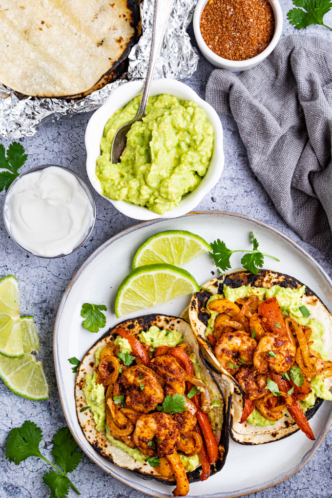 Two shrimp fajitas on a gray plate with limes. A bowl of guacamole, sour cream, and toasted tortilla shells are in the background.