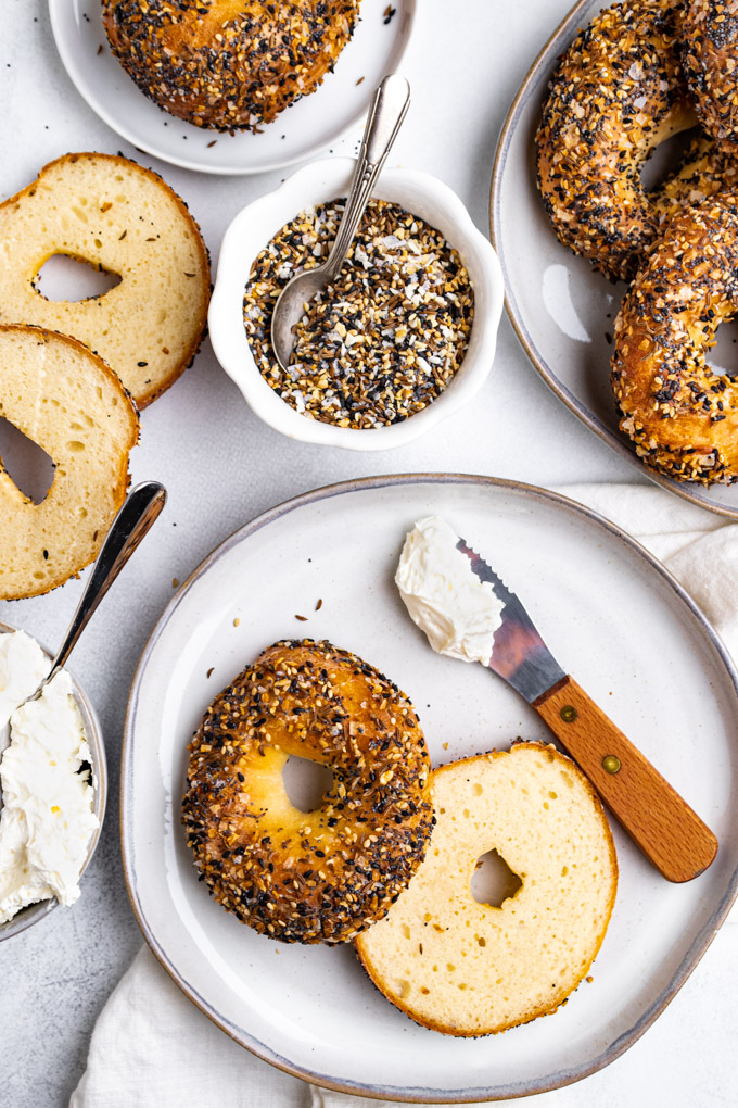 A sliced Greek yogurt bagel on a gray plate, with a knife that has cream cheese on it. Everything bagel seasoning is in a bowl, behind it, a plate of bagels is off to the right, along with another bagels sliced in half, and a bowl of cream cheese is to the left of the plate of bagels.