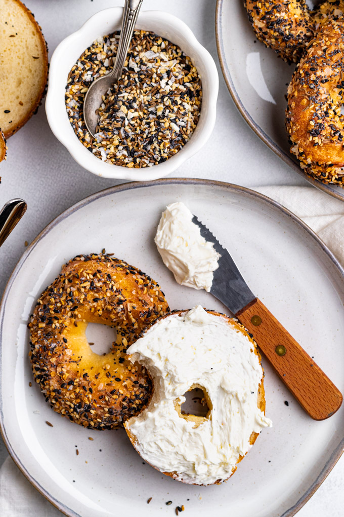 Overhead shot of Greek yogurt bagels. One bagel is sliced in half, on a plate. One half of the bagel is spread with cream cheese, a bite is taken out of that bagel. A small bowl of everything bagel seasoning is behind the bagels, and another plate of bagels is in the corner.