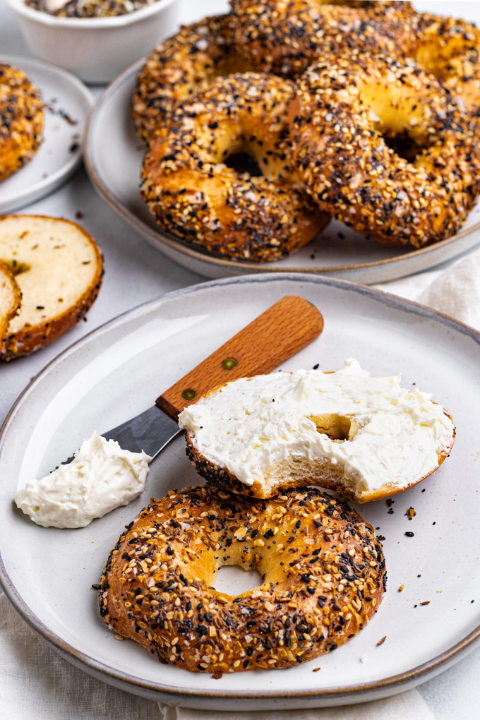 Angled shot of A sliced Greek yogurt bagel on a gray plate, with a knife with cream cheese. Once half of the bagel has cream cheese spread on it, a bite is taken out of that bagel. Another plate is filled with more bagels.