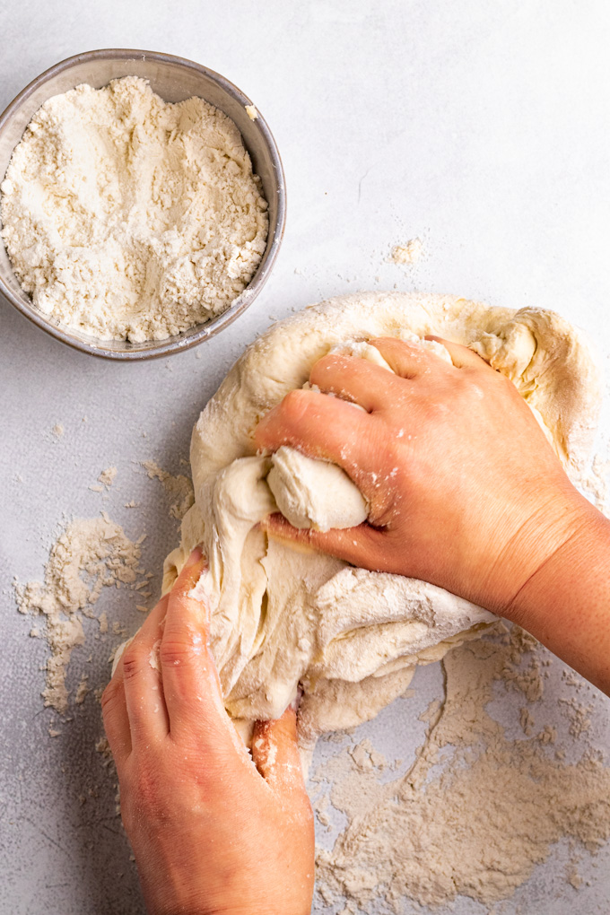 Hands kneading bagel dough, on a light gray background, with a bowl of flour in the corner.