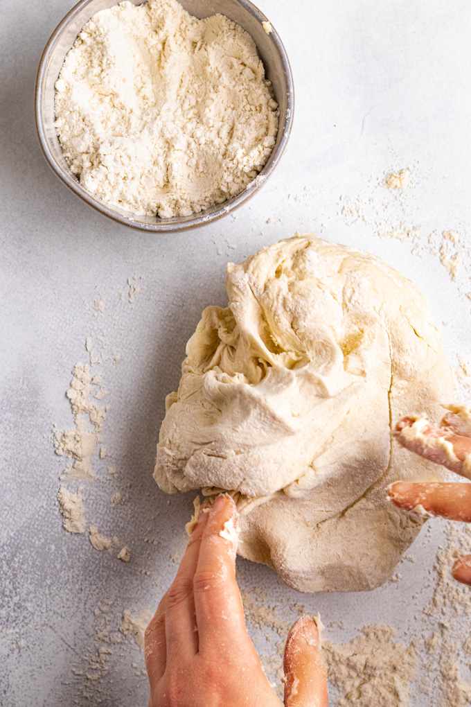 Hands reaching into the corner of the frame, about to knead bagel dough. A bowl of flour is in the corner.