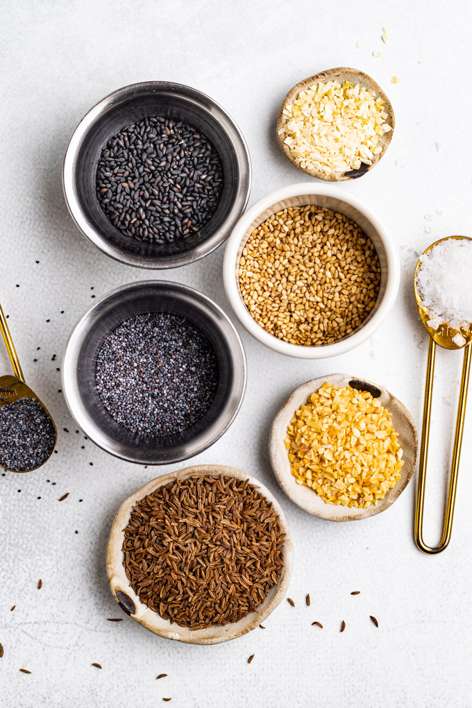 Seeds and spices in separate small bowls: caraway seeds, granulated garlic, sea sat, poppy seeds, white sesame seeds, black sesame seeds, and granulated onion