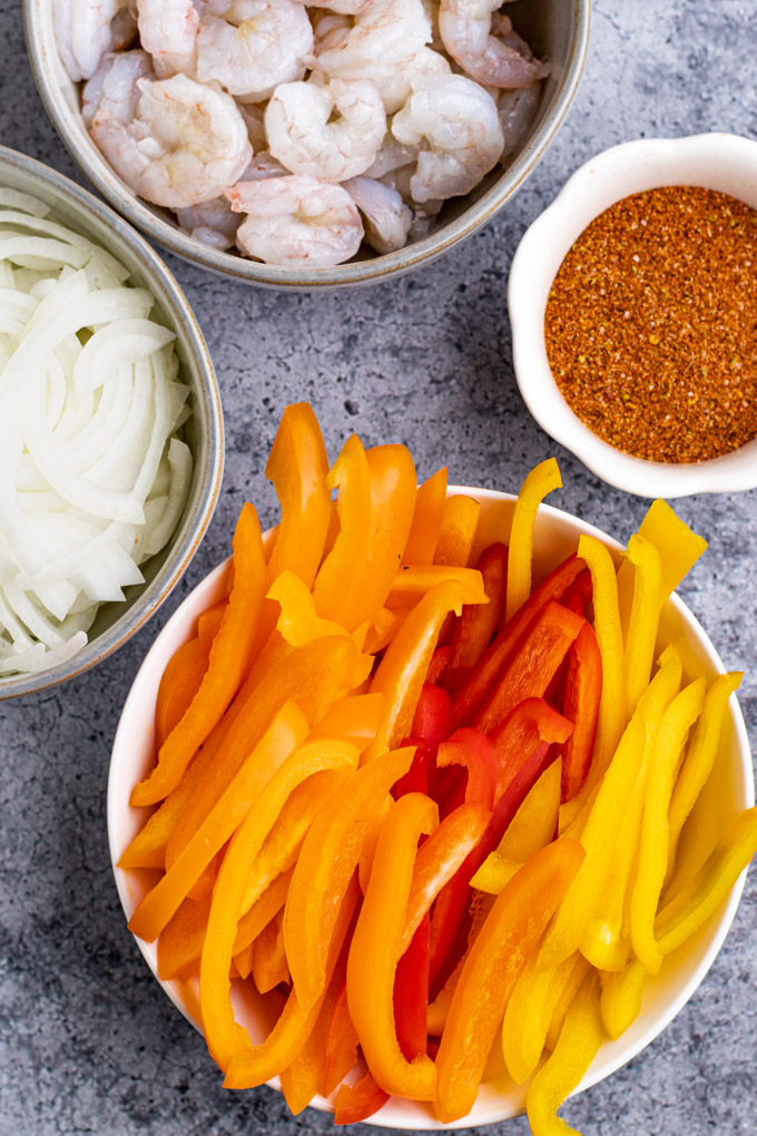 ingredients in separate bowls: bell peppers cut into strips, onion cut into strips, fajita seasoning, shrimp