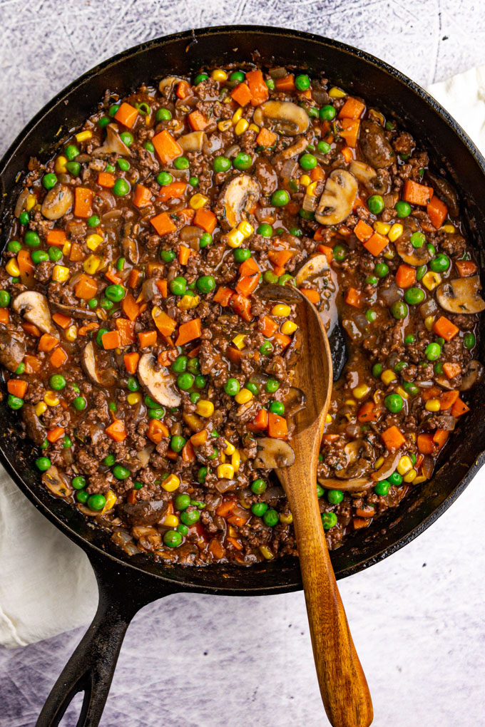 filling for shepherd's pie in a black cast iron skillet with a wooden spoon. There are lots of green peas, mushrooms, carrots, and ground meat in a gravy.