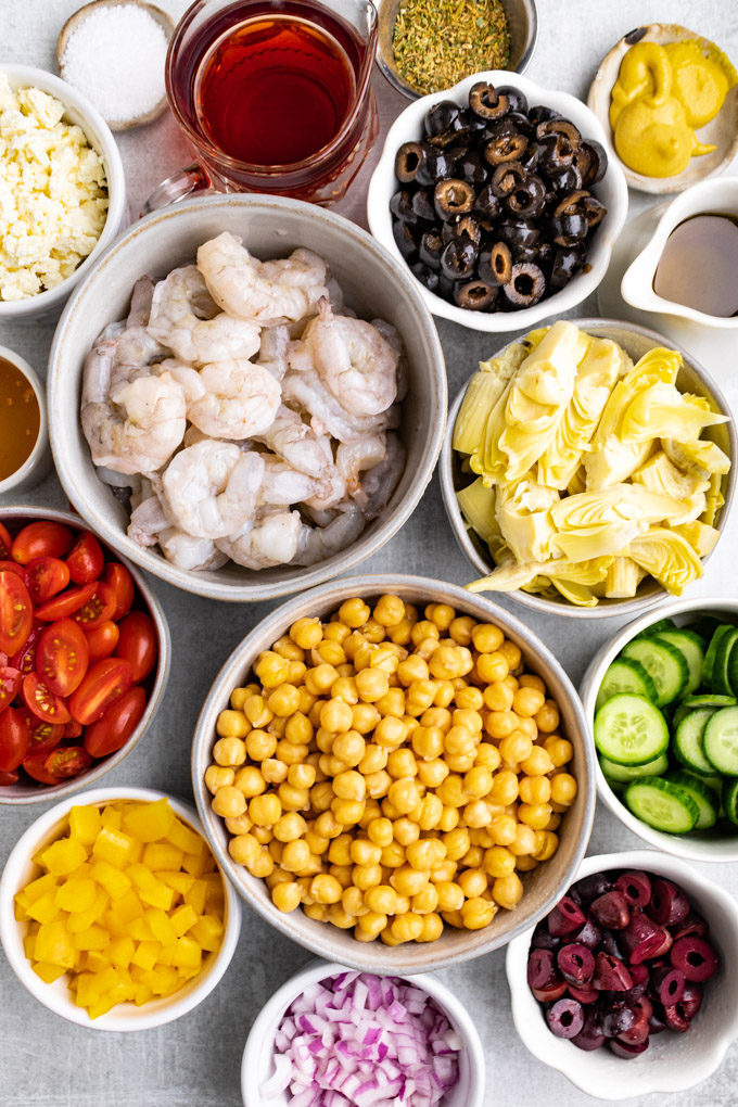 Ingredients for chickpea salad in separate bowls - chickpeas, shrimp, red onion, olives, cucumbers, yellow bell pepper, tomatoes, artichoke hearts, feta cheese etc.