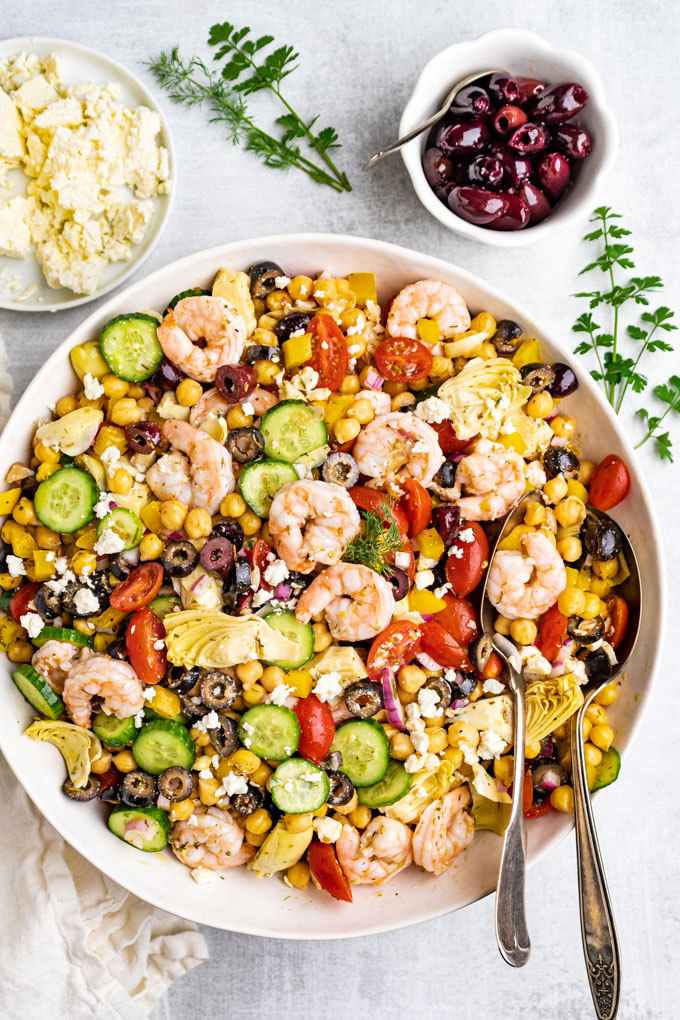 Greek chickpea salad with shrimp in a large serving bowl with 2 serving spoons in it. A bowl of olives are in the background along with a plate of feta cheese.