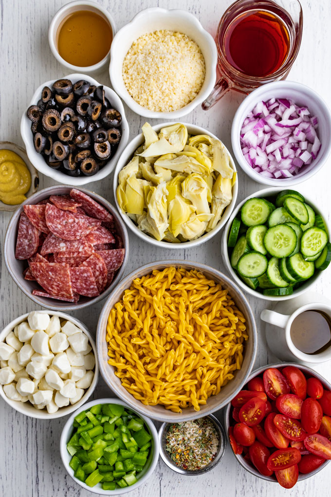 Ingredients are portioned out into individual bowls: dried pasta, salami, olives, artichoke hearts, mozzarella cheese, cucumbers, green bell pepper, tomatoes, mustard, black olives, red onion, parmesan cheese, olive oil, red wine vinegar.