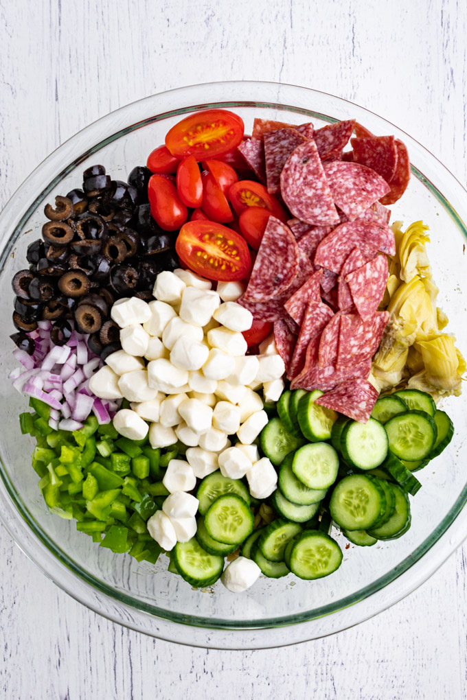 Ingredients for Italian pasta salad in a mixing bowl. Sections of cucumbers, Mozzarella cheese, salami, green bell pepper, tomatoes, and black olives.
