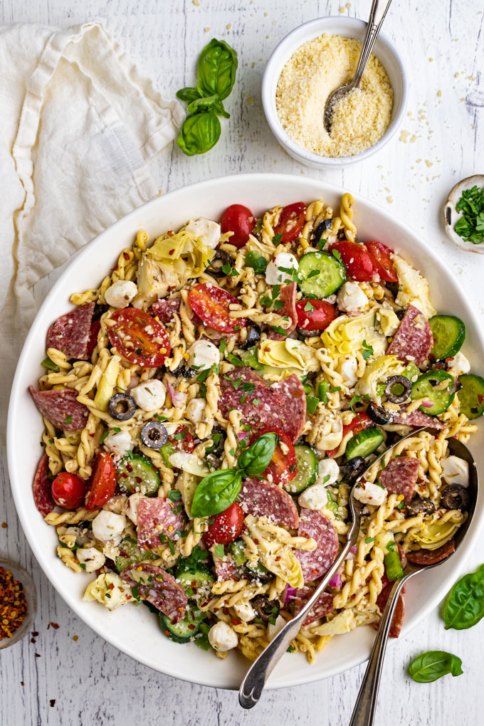 Italian pasta salad with salami in a larege white serving bowl. 2 serving spoons are in the salad. A napkin is off to the left, with a bowl of parmesan cheese that has a spoon in it. A small bowl of parsley, and a small bowl of red pepper flakes are on either side of the bowl.