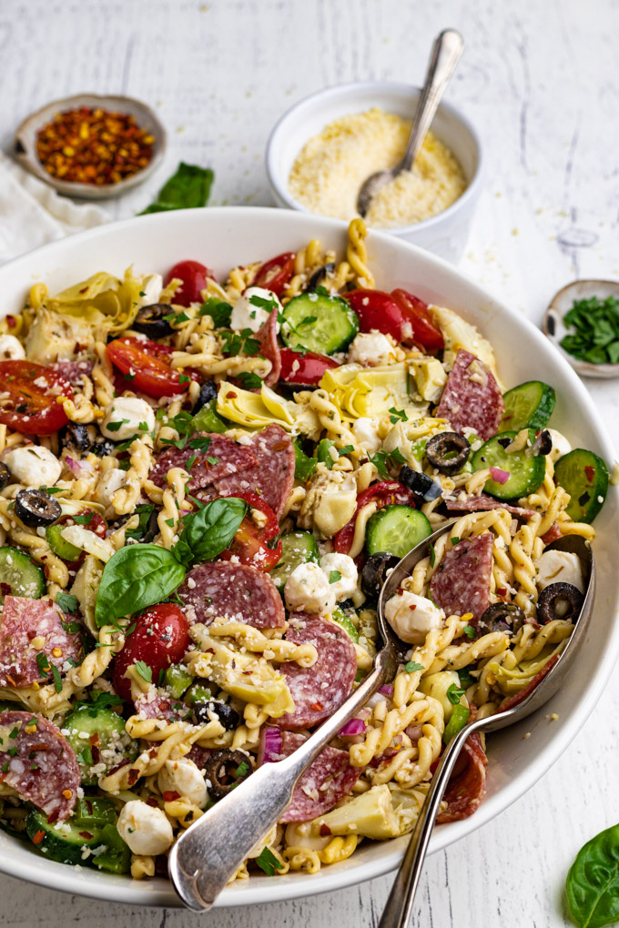 An angled, close up shot of Italian pasta salad with salami. Serving spoons are in the pasta salad, and a bowl of parmesan cheese is in the background, along with red pepper flakes.