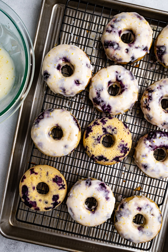blueberry donuts are on a a wire rack. Most of them are glazed, while a few aren't - a bowl of glaze is off to the side.