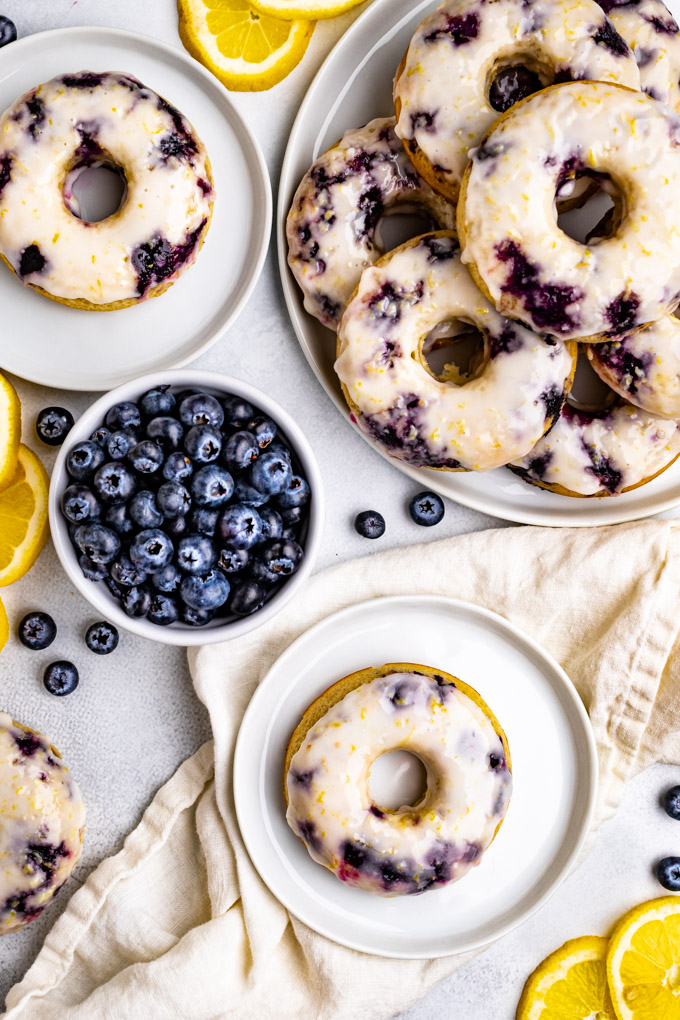 Blueberry cake donuts with lemon glaze. One donut is on a small plate, with a large plate of donuts off to the right, and a bowl of blueberries.