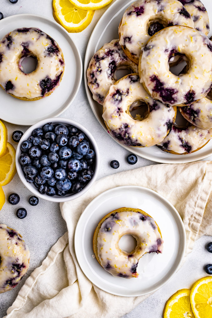 An overhead shot of blueberry cake donuts with lemon glaze. One donut is on a small plate with a bite taken out of it. There is a small bowl of blueberries, and a larger plate with a stack of more donuts.
