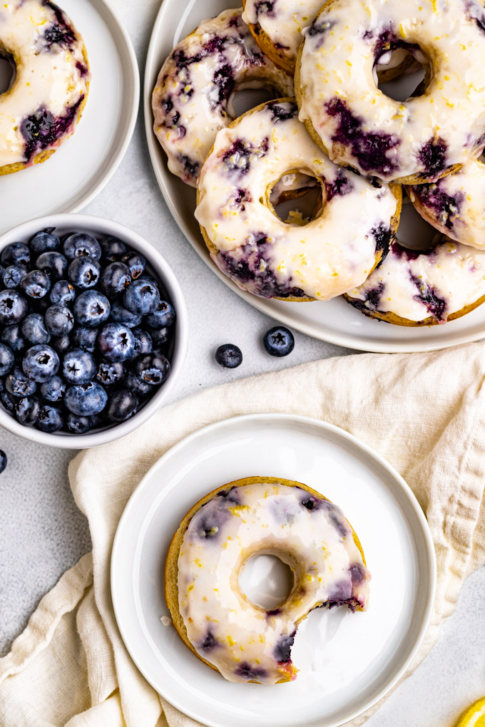 Overhead shot of a blueberry bake donut with a bite taken out of it. A larger plate is stacked high with more donuts, along with a small bowl of blueberries.