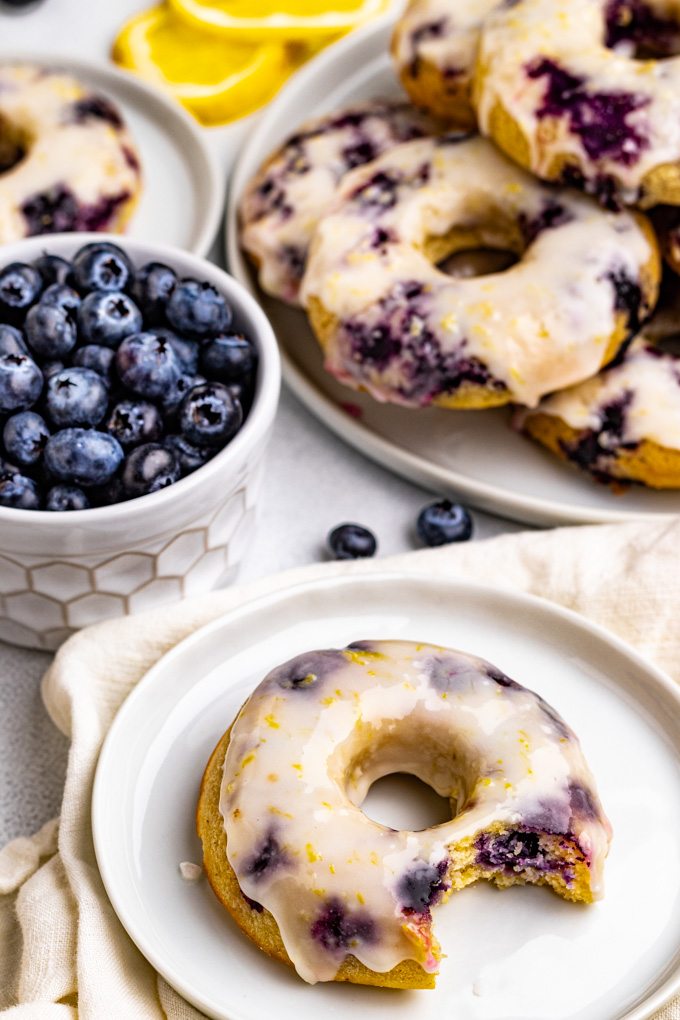 A closeup angled shot of blueberry cake donuts with lemon glaze. One donut is on the plate with a bite taken out of it. A small bowl of blueberries is behind it, and a larger plate with donuts stacked high is in the background.