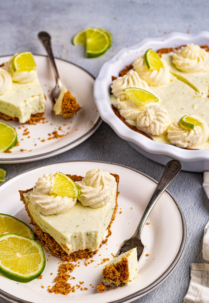 Two slices of the best key lime pie on plates with a bite taken out of them. An angled, close up shot. The pie plate with the remaining pie is in the background.. There are dollops of whipped cream, and slices of lime decorating the pie.