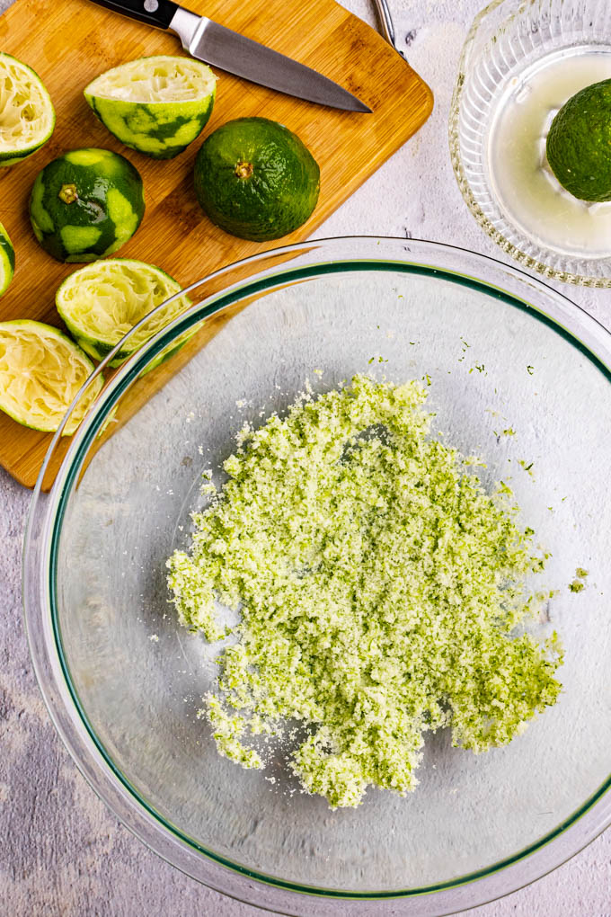 Sugar and lime zest rubbed together in a bowl to make lime sugar. Juiced, and zested limes are in the background, on a cutting board.