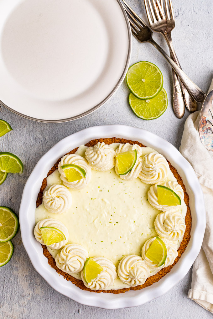 Overhead shot of the best key lime pie with rosettes of whipped cream decorating the outside, and lime slices on every other rosette. There are plain plates, a pie server, and forks in the background.
