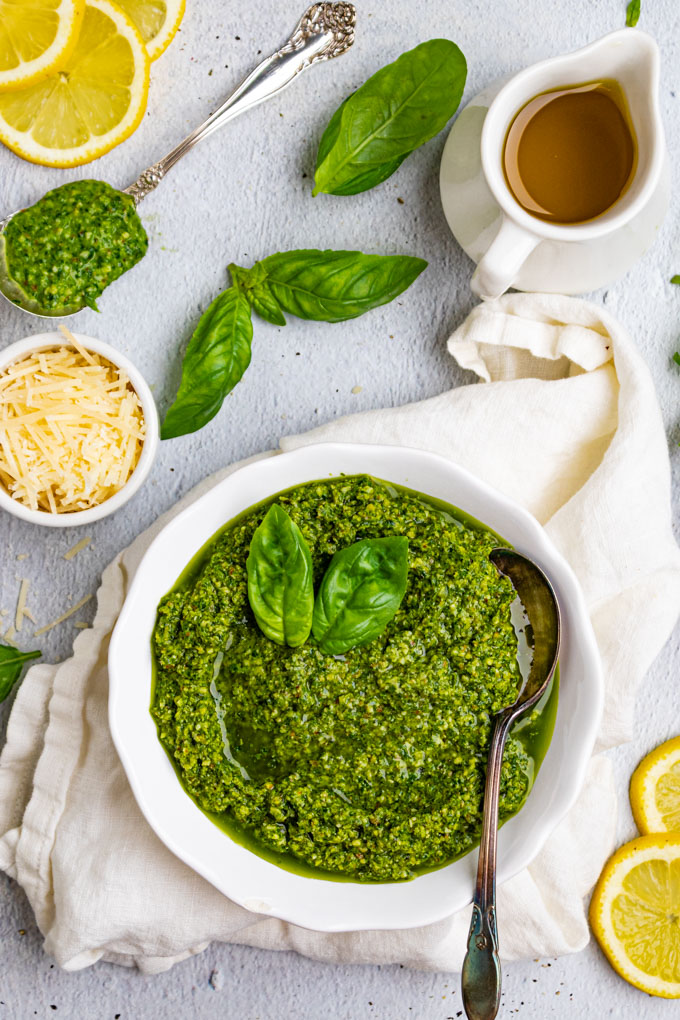 basil pesto recipe in a white bowl with a spoon. Basil leaves, and lemon slices are scattered in the background. Along with a bowl of parmesan cheese, and a spoonful of pesto.