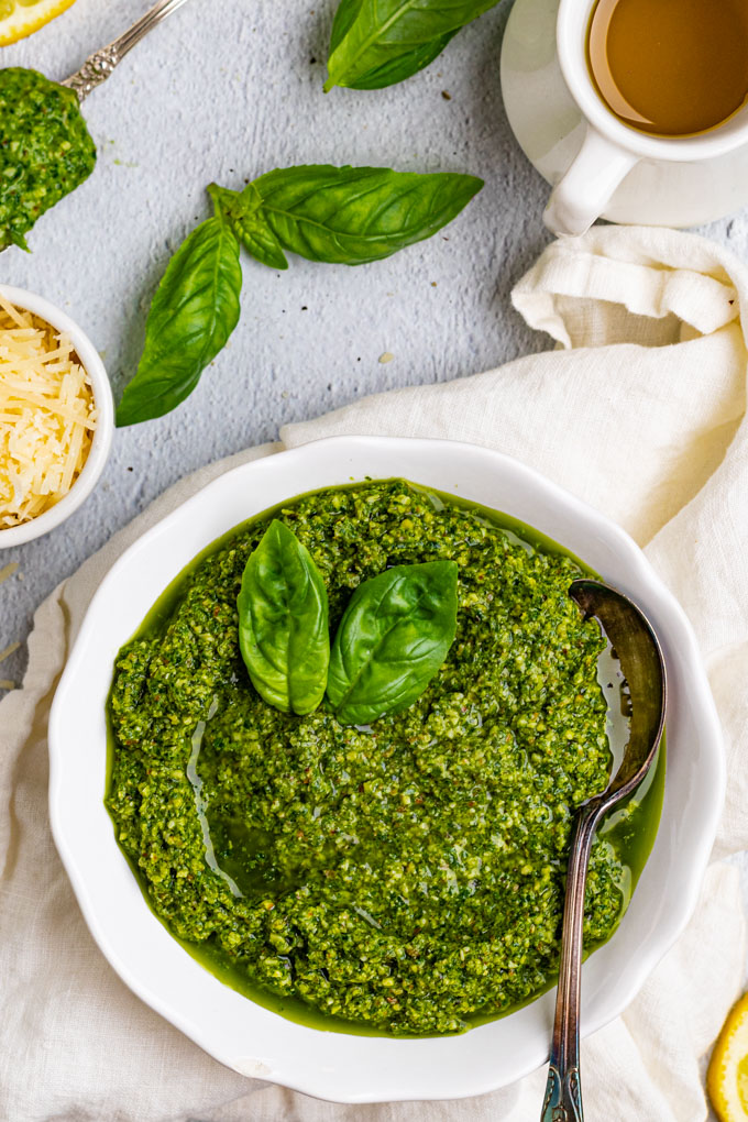 Overhead shot of basil pesto recipe in a white bowl with a spoon. There are a few basil leaves scattered, and a jug of olive oil in the background.
