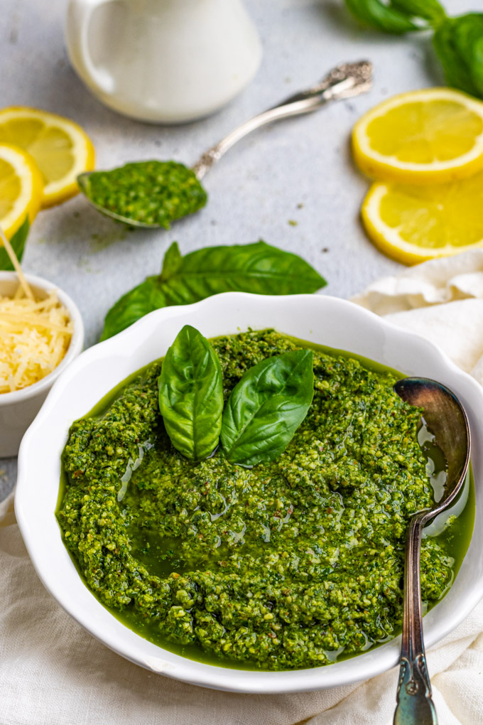closeup angled shot of basil pesto recipe. Pesto is in a white bowl with lemon slices in the background.