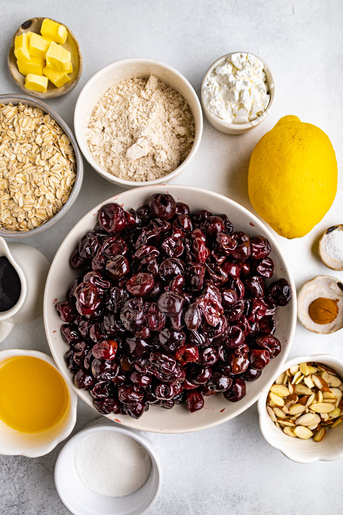 Ingredients for cherry crisp are in bowls - frozen cherries, sliced almond, sugar, butter, oats, maple syrup, oat flour, cornstarch, lemon