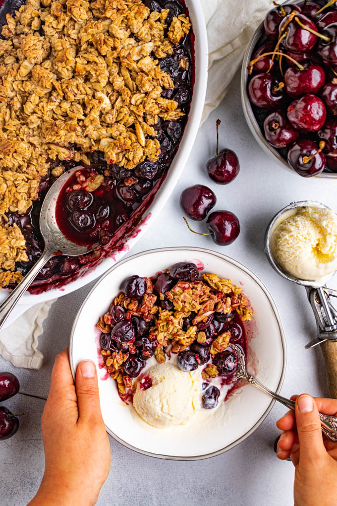 Cherry crisp with frozen cherries. The cherry crisp is in a bowl with a scoop of vanilla ice cream. A hand is holding a spoon and another hand is holding the bowl. Fresh cherries are in a bowl in the background, and scattered around. A scoop of ice cream is off to the side. And a the baking dish filled with cherry crisp is off to the side.