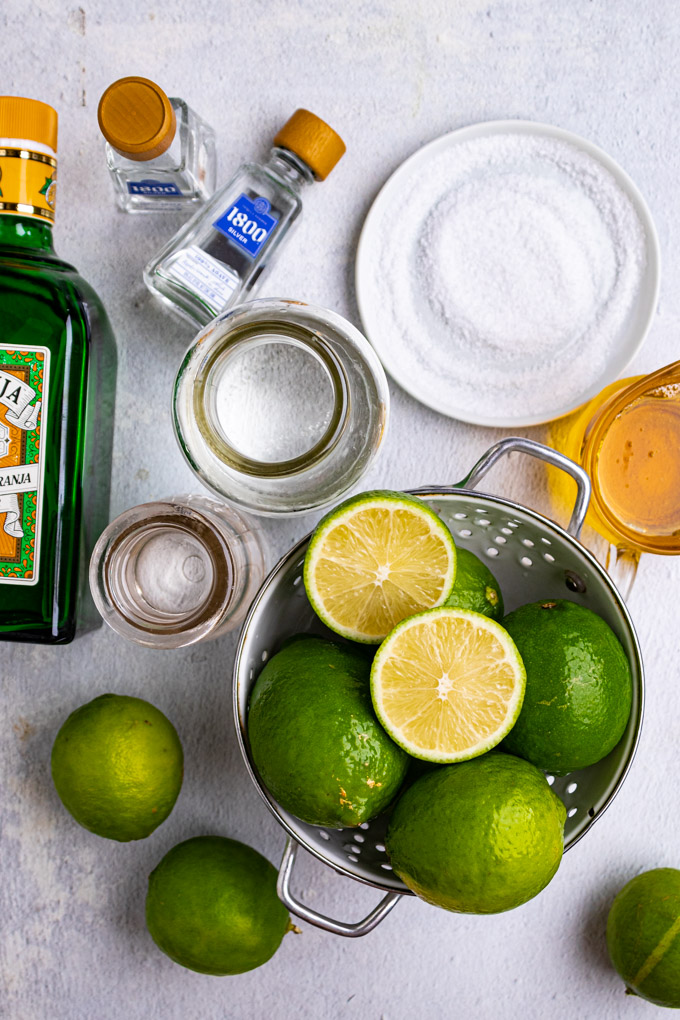 Overhead shot of ingredients for margaritas: limes, agave nectar, plate of salt, tequila, and orange liqueur.