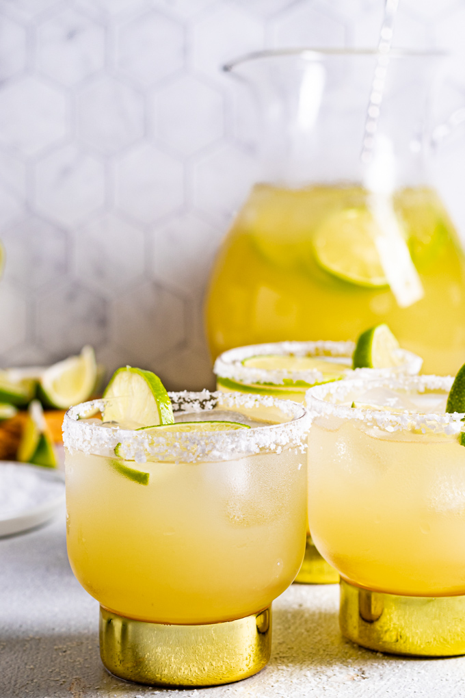 A close up of pitcher margaritas, straight on. 3 glasses filled with margaritas and a salted rim are in the front. A pitcher of margaritas is in the back, to the left in the background are lime wedges on a cutting board.