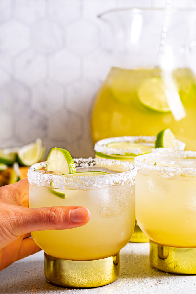 Close up shot of pitcher margaritas. A hand is holding one of the 3 glasses of margaritas. A pitcher is in the background filled with margarita.