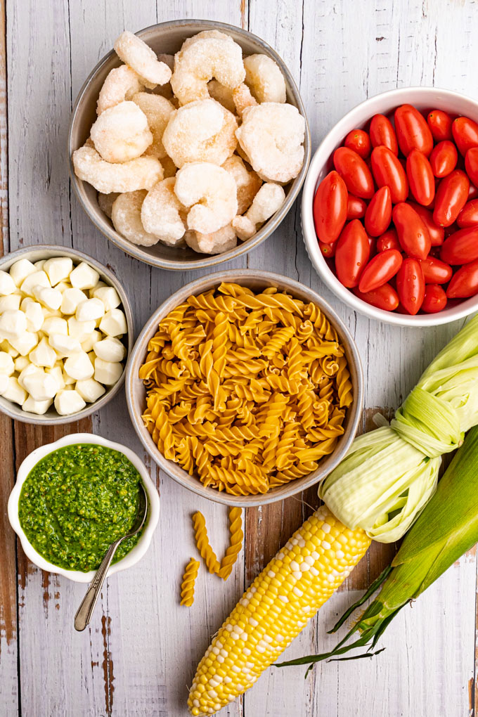 Ingredients in bowls, for pesto pasta salad. Pasta, tomatoes, pesto, mozzarella cheese, shrimp, and corn on the cob.
