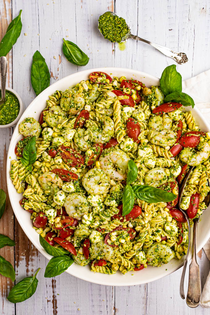 Pesto pasta salad with shrimp in a serving bowl, on a white wood background with basil leaves scattered around.
