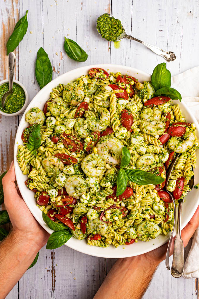 Pesto pasta salad with shrimp, in a large serving bowl with hands holding the bowl. There are basil leaves scattered in the background.